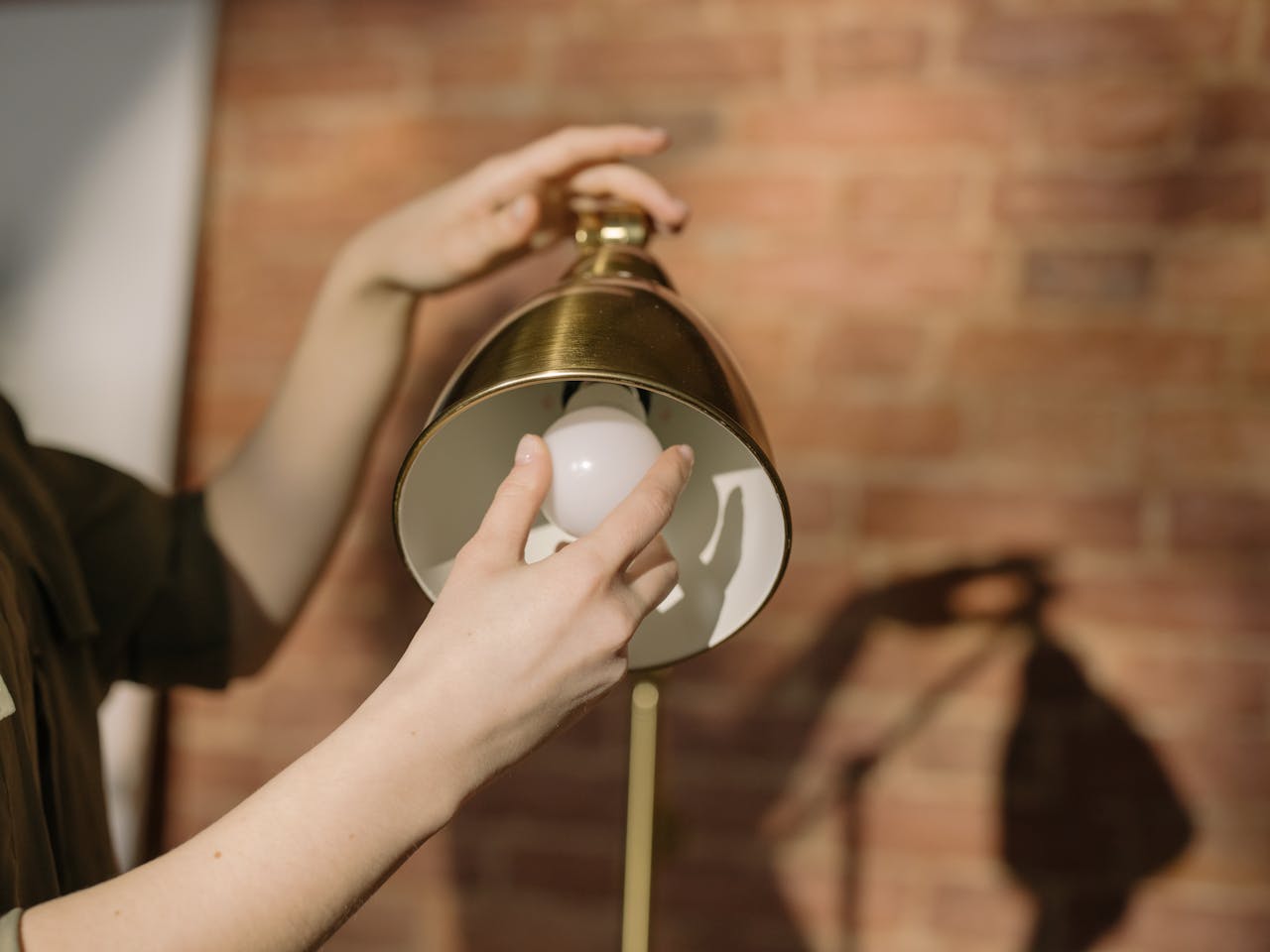 Hands changing a light bulb in a lamp with a brick wall background.